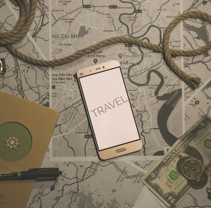 A flat lay composition featuring a smartphone with the word 'TRAVEL' displayed on its screen, positioned on top of several maps. A coiled piece of rope lies across the scene, accompanied by a wristwatch, a notebook with a pen, and some currency including paper bills and coins. The overall theme suggests preparation or planning for a journey.