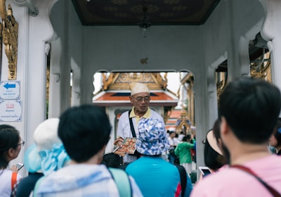 A group of tourists are gathered around a guide who is holding a brochure or map. The setting appears to be a historical or cultural site with ornate architecture in the background and an archway. The guide is wearing a hat and glasses, and the tourists are mostly wearing casual clothing and hats.
