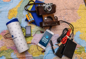 A collection of travel essentials sits on a detailed map. A vintage camera in a brown leather case rests near a smartphone displaying Cyrillic text. Next to it, a large white travel tumbler marked 'Journal' stands beside several electronic gadgets, including a portable music player, an external hard drive, and a set of coiled cables. A pack of color film and a blue notebook complete the setup.