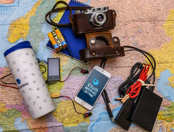 A collection of travel essentials sits on a detailed map. A vintage camera in a brown leather case rests near a smartphone displaying Cyrillic text. Next to it, a large white travel tumbler marked 'Journal' stands beside several electronic gadgets, including a portable music player, an external hard drive, and a set of coiled cables. A pack of color film and a blue notebook complete the setup.